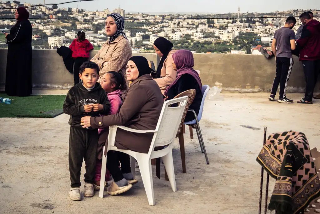 Several Palestinian neighbors stand closely together in Tarqiyah, West Bank, watching as Israeli military machinery demolishes a residential house on February 1, 2026; dust and debris fill the air, conveying the shock and shared impact on the surrounding community.
