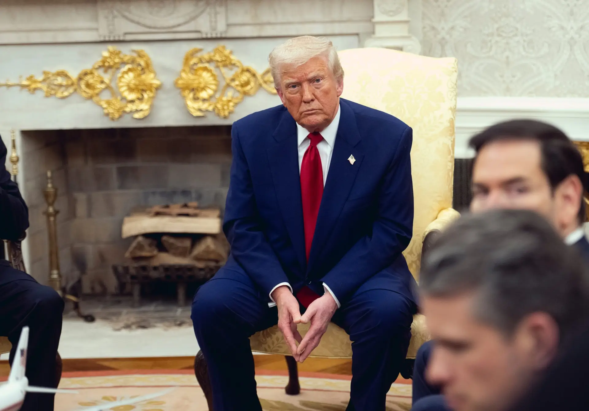 President Donald Trump speaking to reporters in the Oval Office at the White House, seated behind the Resolute Desk with U.S. flags in the background