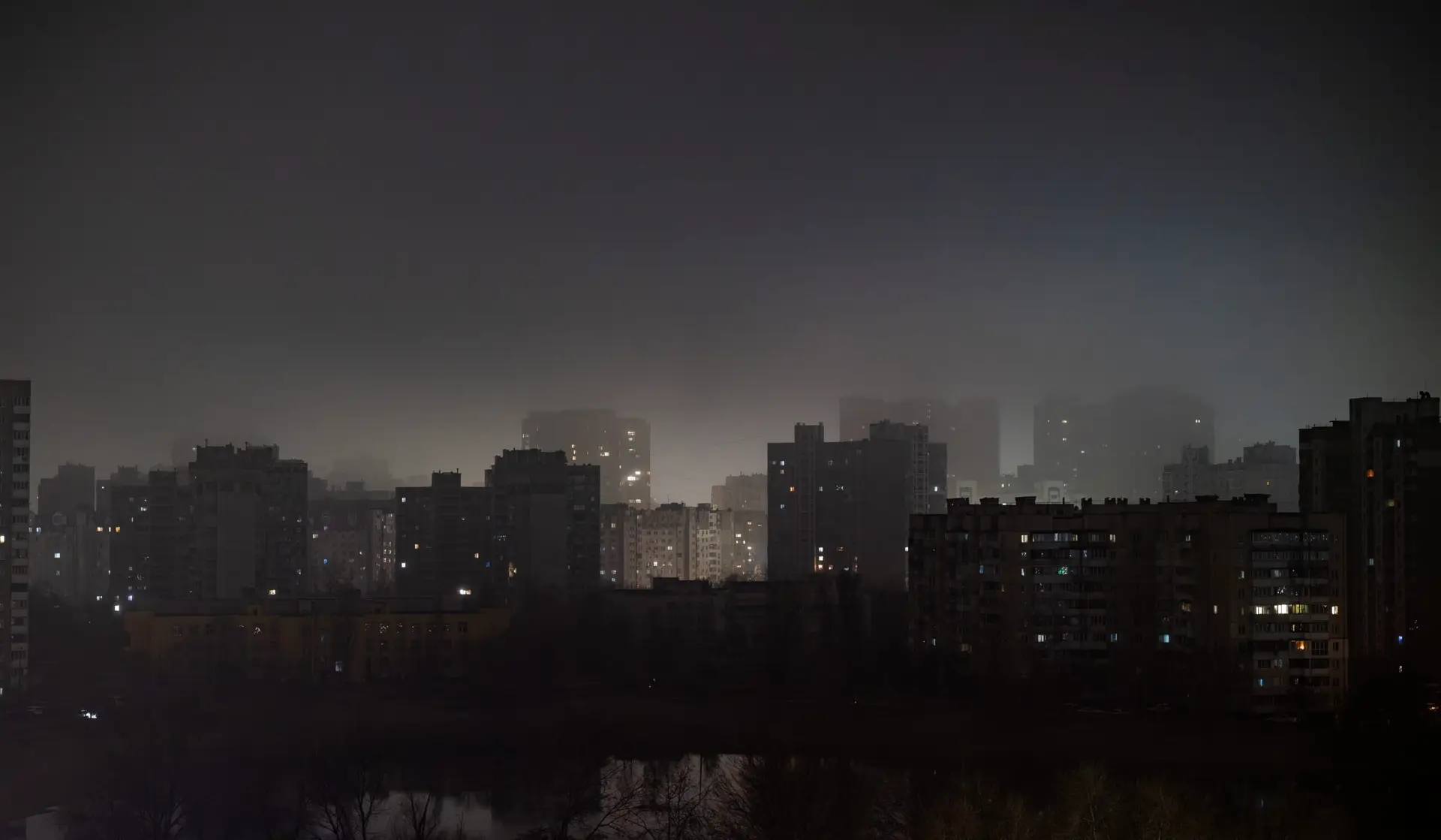 Kyiv, Ukraine at night during a total blackout in winter 2024, with dark silhouettes of high-rise residential buildings around a lake and only a few scattered lights visible, illustrating the impact of Russian attacks on the power grid and the vulnerability of civilian urban life in wartime.